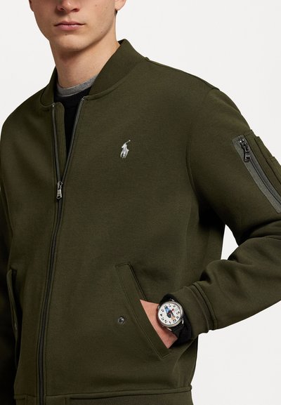 Casaco bomber verde-oliva com fecho frontal de correr, gola canelada e bolso com fecho de correr na manga; apresenta um pequeno logotipo bordado. Relógio preto visível.