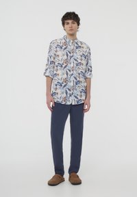 Camicia a maniche corte floreale di colore bianco con motivi blu e marroni; abbinata a pantaloni blu navy e scarpe slip-on marroni. Design della colletto semplice.