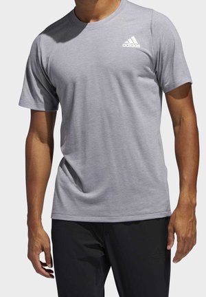 Mann trägt ein hellgraues Adidas-T-Shirt mit kurzen Ärmeln und schwarze Adidas-Hose, steht vor einem einfarbigen Hintergrund.