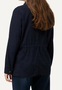 Marinblå lättviktsjacka med långa ärmar, texturerat tyg, elastisk midja och knappdetaljer vid ärmslut.