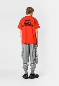 Camiseta roja con texto bordado en negro "MOD WAVE MOVEMENT" en la parte posterior, combinada con pantalones cargo grises y zapatos negros sin cordones.