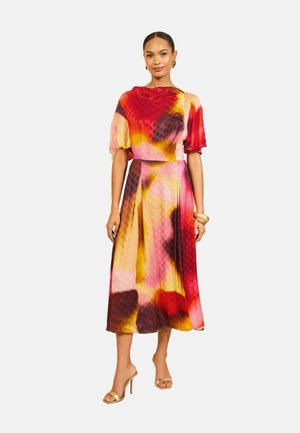 Frau trägt ein farbenfrohes Midikleid mit roten, gelben, pinken und schwarzen abstrakten Mustern, kurzen Ärmeln und goldenen Sandalen mit hohen Absätzen.