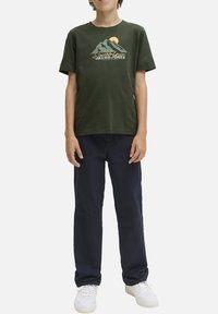 T-shirt en coton vert avec un graphisme de montagne et de soleil. Associé à un pantalon marine et des baskets blanches, se tenant devant un fond uni.