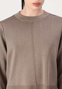 Brązowy sweter z prążkowanego materiału, z wysokim dekoltem, z centralnym szwem z przodu i długimi rękawami, wykonany z miękkiego, teksturowanego materiału.