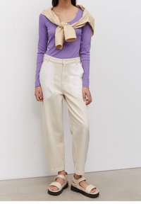 Lila långärmad topp, beige byxor med vita fläckar och beige sandaler med tjocka svarta sulor. Slät textur och avslappnad passform.