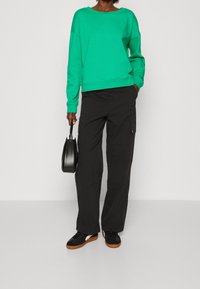 Sudadera verde con un cuello ancho, combinada con pantalones cargo negros. Bolso negro y zapatillas negras con suela marrón. Diseño simple, ajuste casual.