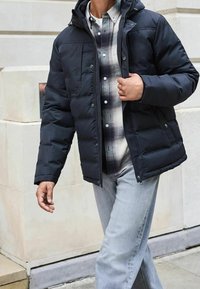 Schwarze Steppjacke mit Kapuze, vorne mit Reißverschluss und Knopfdetails sowie zwei Fronttaschen, getragen über einem karierten Hemd und kombiniert mit hellen Jeans.
