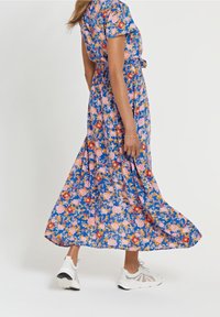 Blumenkleid im Maxistil mit kurzen Ärmeln, auf blauem Hintergrund mit pinken/roten Blumen. Kombiniert mit weißen Sportschuhen für einen Kontrast.