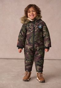 Zaļi un brūni ierocēto rakstu pūkainais sniega kostīms ar mākslīgās kažokādas kapuci, priekšējo rāvējslēdzēju un melnām aprocēm. Brūnas kurpes ar auklēm noslēdz izskatu.