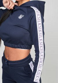 Žena v námořnickém modrém zkráceném hoodié a odpovídajících teplácích s bílým logem a textem "SIKSILK" podél rukávů a kapes kalhot.