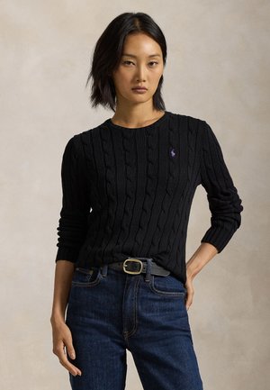 Young woman wearing a black cable-knit sweater with purple logo, dark blue jeans, and a black belt, posing against a neutral background.
