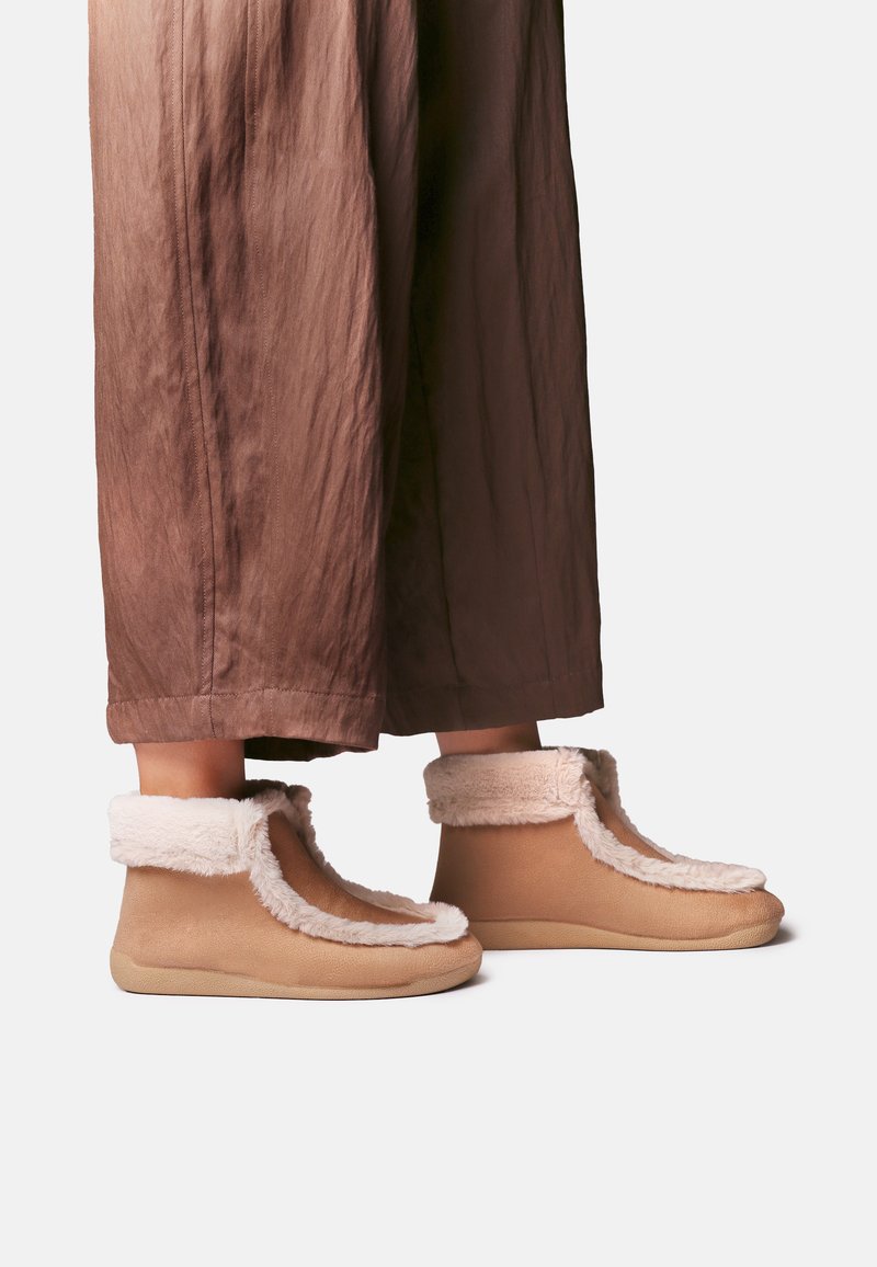 Pieds portant des chaussons d'intérieur marron clair avec une doublure en fourrure blanche, associés à un pantalon marron ample sur un fond clair uni.