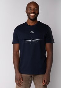 T-shirt en coton bleu marine avec un design graphique représentant une silhouette se tenant près d'une ligne d'eau, sous un grand corps céleste, imprimé en blanc.