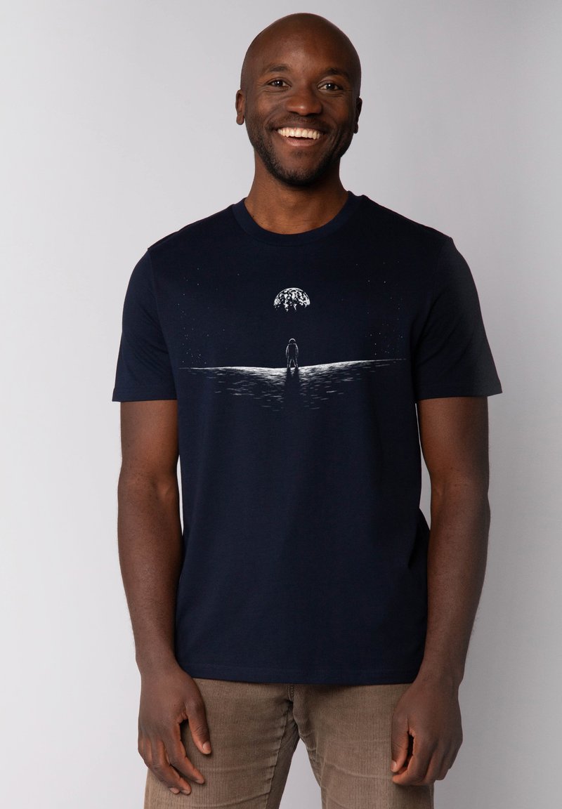 T-shirt en coton bleu marine avec un design graphique représentant une silhouette se tenant près d'une ligne d'eau, sous un grand corps céleste, imprimé en blanc.