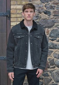 Melnas denima veste ar mākslīgo aitu apmali, pogu aizdari, divām krūšu kabatām un redzamām šuvēm. Modelis valkā baltu krekla apakšā.