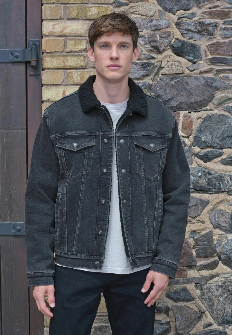 Črna denim jakna z umetnim ovčjim krznom na ovratniku, z gumbi na pritiskanje, dvema žepoma na prsih in vidnimi šivi. Model nosi belo srajco spodaj.