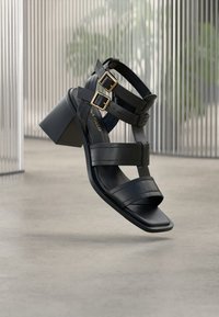 Μαύρα σανδάλια με τακούνι που διαθέτουν πολλά ρυθμιζόμενα λουριά, χρυσές αγκράφες και χοντρό τακούνι. Λείο υλικό με μινιμαλιστικό σχεδιασμό.