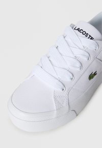 Valge lõuendist vabaajajalats tekstureeritud pinnaga, ümmargune nina ja kummitald. Varustatud valgete paeltega ja rohelise Lacoste logoga, mis on küljele tikitud.