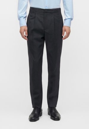 Mężczyzna ubrany w czarne, dopasowane spodnie, jasnoniebieską elegancką koszulę i czarne eleganckie buty, stojący na tle jednolitego białego tła.