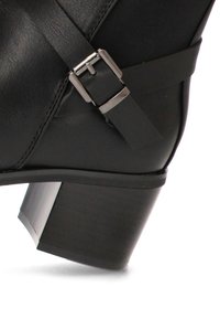 Bota negra de tacón con parte superior de cuero liso, una correa con un hebilla metálica y un tacón cuadrado con superficie texturizada.