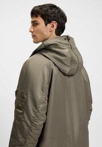 Khaki pufferjack met capuchon, met een gestructureerde afwerking, zijzakken met rits en een logo-accent op de mouw.