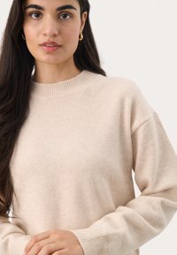 Pull beige en mailles douces ; encolure ronde avec bordure côtelée, épaules tombantes et manches longues. Texture lisse avec des accents minimalistes.