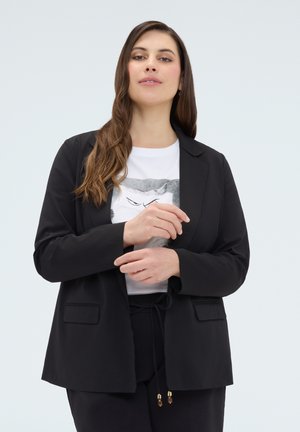 Vrouw met lang bruin haar die een zwarte blazer draagt over een wit T-shirt met een kattengezicht, ze past de mouwen van haar blazer aan.