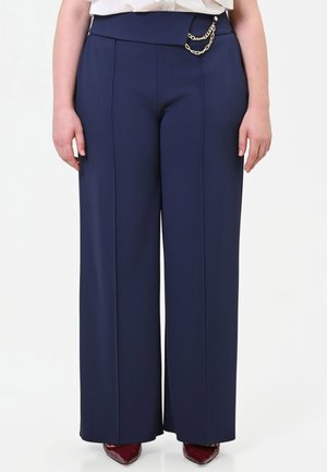 Pantaloni a gamba larga blu navy con cuciture frontali e dettaglio di catena dorata in vita, indossati con scarpe appuntite bordeaux.