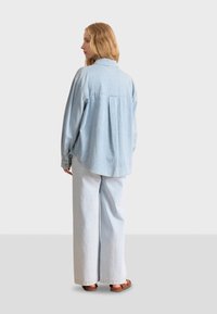 Lichtblauwe spijkerblouse met lange mouwen, een gebogen zoom en een knoopenkraag, gecombineerd met lichtblauwe brede broek en bruine sandalen.