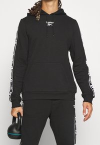 Svart huvtröja i bomullsblandning, med kenguruficka, dragskohuva och vita geometriska detaljer på ärmarna.