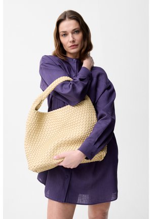 Vrouw in een paarse jurk met knopen, die een grote beige geweven handtas vasthoudt tegen een effen lichte achtergrond.