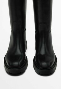 Bottes courtes en cuir noir avec une semelle épaisse et une texture lisse. Elles présentent un bout arrondi et des accents cousus discrets sur les côtés.