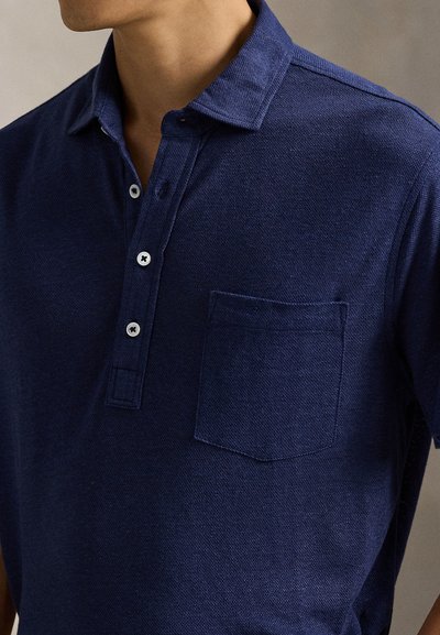 Πόλο μπλούζα σε ναυτικό μπλε χρώμα κατασκευασμένη από υφασμάτινη υφή, με κουμπωτό πατιλέτα, κοντά μανίκια και τσέπη στο στήθος με ίσια ραφή στο κάτω μέρος.