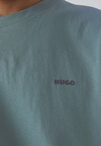 Gaišs teļa krekls, izgatavots no mīksta kokvilnas, ar apaļu kaklu un mazu, tumši zilu "HUGO" logo uz krūtīm. Gluda tekstūra, ikdienas dizains.