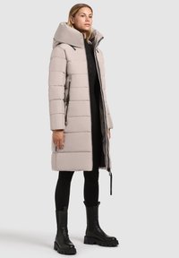 Lys beige pufferjakke med høj krave, lodret quiltning, frontlynlås og sidelommer. Bæres over sort tøj og robuste sorte støvler.