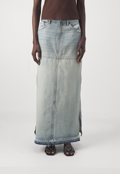 Denim maxi rok in lichtblauw, met verwerkte details, zijsplits en een ruwe zoom. Gepaarde met zwarte open teen sandalen.