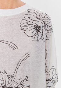Vit genomskinlig topp med svarta blommönster och text. Designen inkluderar stora blomtryck med fina detaljer och en avslappnad passform.