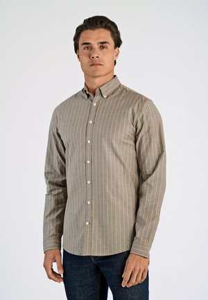 Mann trägt beige, vertikal gestreiftes Hemd mit Knöpfen und dunkelblaue Jeans, steht vor einem schlichten hellen Hintergrund, blickt nach vorne.