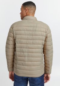 Pufferjakke i lysebeige, quiltet design med horisontale sømme, høj krave og lange ærmer. Båret over mørke denimjeans.