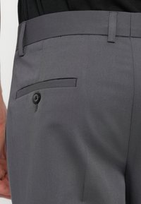 Gray tailored trousers feature a smooth fabric, flat front, and a back pocket with a button closure. Subtle stitching details enhance the design.
