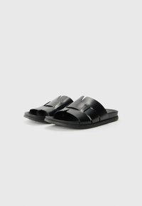 Black slide sandals feature a smooth, matte finish with wide, overlapping straps and a flat sole. The design emphasizes simplicity and comfort.