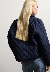 Giacca navy trapuntata con una forma squadrata, caratterizzata da motivi a rombi e polsini elastici, abbinata a jeans blu a vita alta.