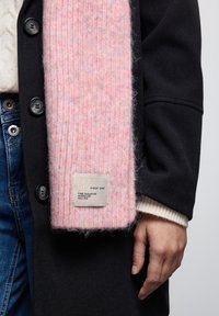 Sciarpa rosa in maglia con un motivo testurizzato, dotata di un'etichetta cucita che recita "BARA STREET ONE." Indossata sopra un cappotto nero con bottoni.