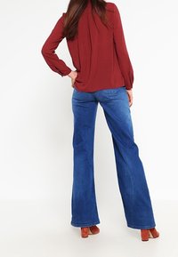 Blouse rouge texturée à manches longues, associée à un jean évasé bleu. Le jean a une finition classique en denim avec un léger délavage, complété par des talons marron.