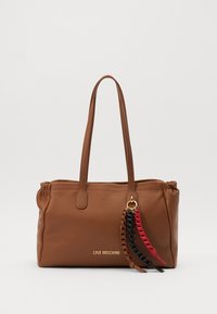 Brown leather tote bag with textured finish, double handles, and a decorative charm featuring red, black, and braided accents. Gold logo visible.