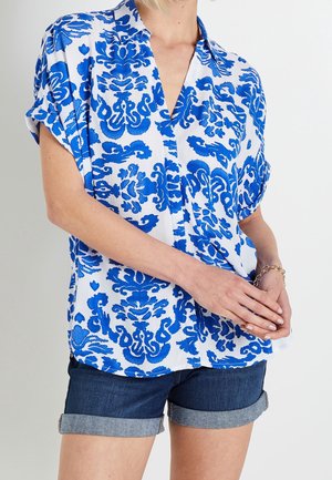 Donna che indossa una camicetta bianca a maniche corte con motivi floreali blu e pantaloncini di jeans scuri arrotolati, con le mani giunte davanti.