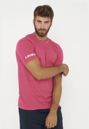 Homme avec barbe portant un t-shirt rose LEGEA ajustant sa manche gauche, debout sur un fond blanc uni.