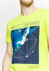 Neon žluté bavlněné tričko s grafickým designem obsahujícím modré a černé části, geometrické liniemi a textem o ultrafialovém světle.