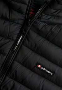 Veste doudoune noire avec texture matelassée, dotée d'une fermeture éclair noire et d'un petit patch logo "Superdry" rouge et blanc sur la poitrine.