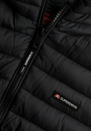 Chaqueta de plumas negra con textura acolchada, que presenta una cremallera negra y un pequeño parche con el logo "Superdry" en rojo y blanco en el pecho.